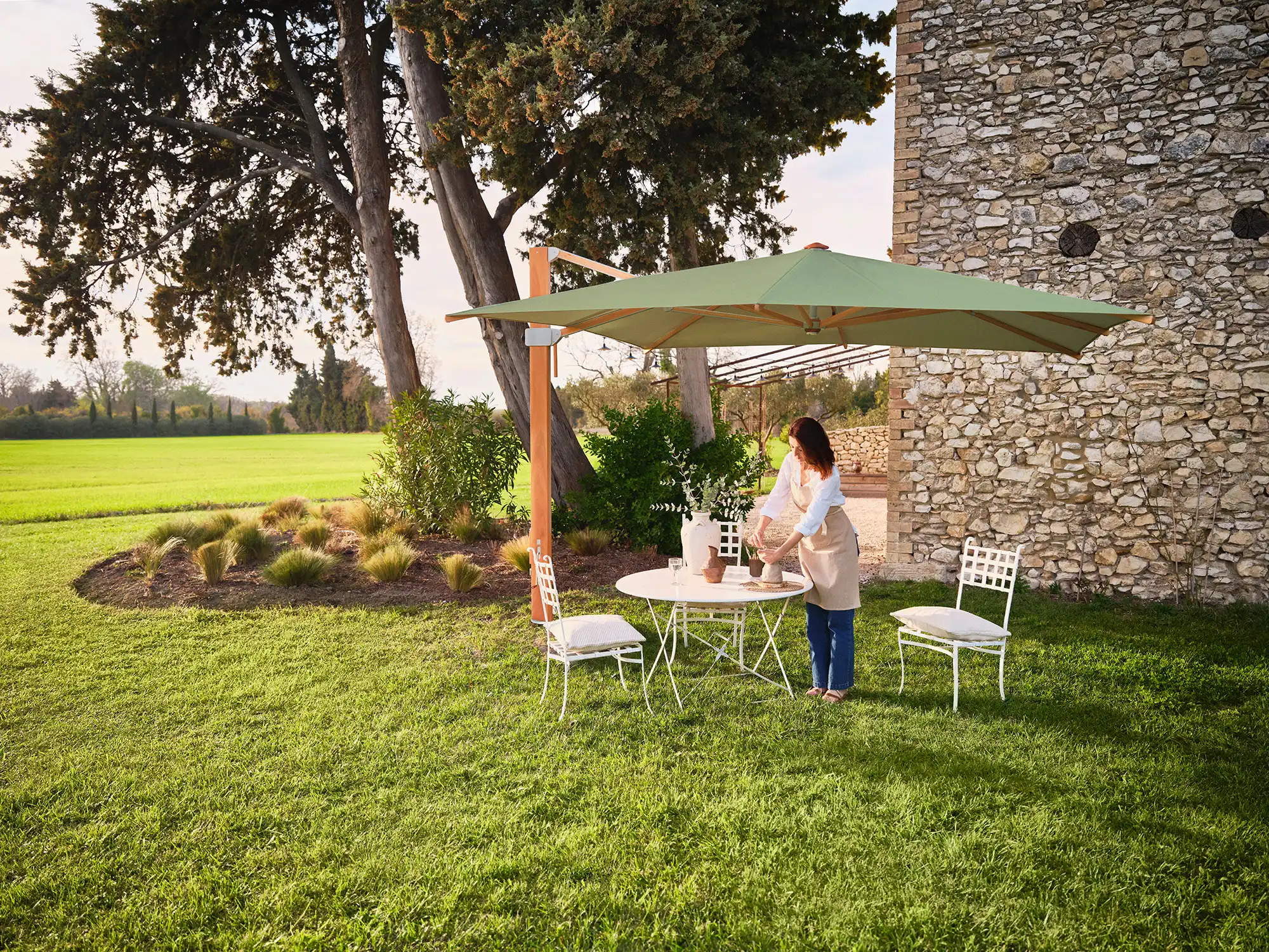 Vor einem alten gemauerten Gebäude im Schatten eines Baumes ist ein gemütlicher Essbereich mit Metallgeartenmöbeln, geschützt durch einen grünen Sonnenschirm