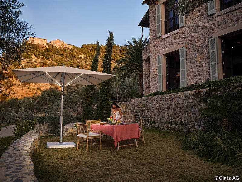 Großer Sonnenschirm Glatz Fortello auf einer Terrasse vor einem Natursteinhaus in warmer Abendstimmung