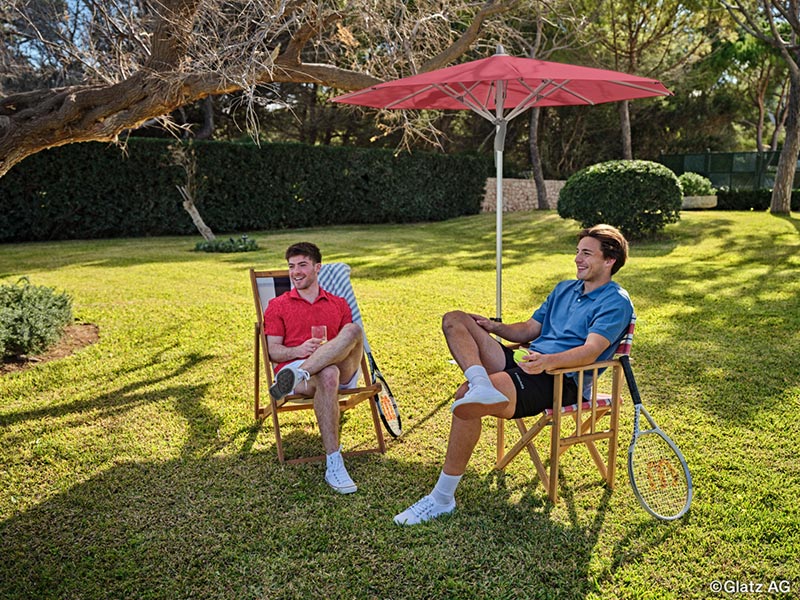 Zwei Männer entspannen im Garten unter einem roten Sonnenschirm Glatz Fortino auf einer grünen Rasenfläche