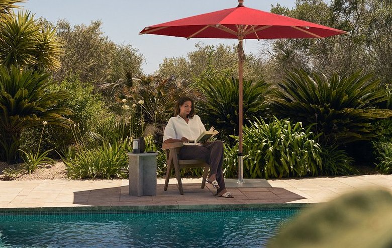Eine Frau sitzt am Pool auf einem Stuhl geschützt unter einem Sonnenschirm