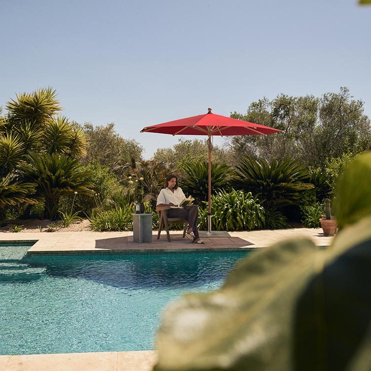 ein Pool und imHintergrund sitzt eine Frau im Schutz eines roten Sonnenschirmes